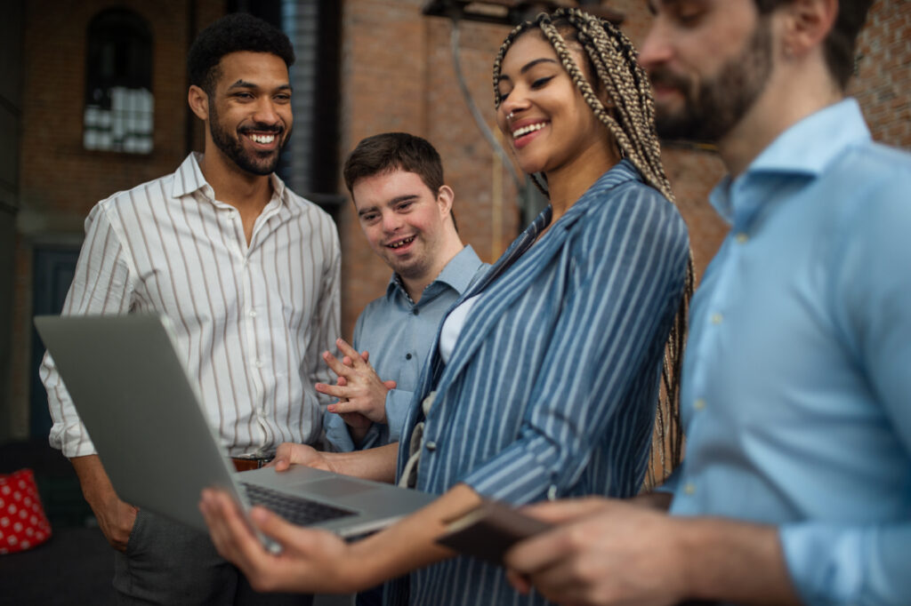 A small group stands outdoors near a brick building, collaborating around an open laptop and a mobile phone during a discussion.