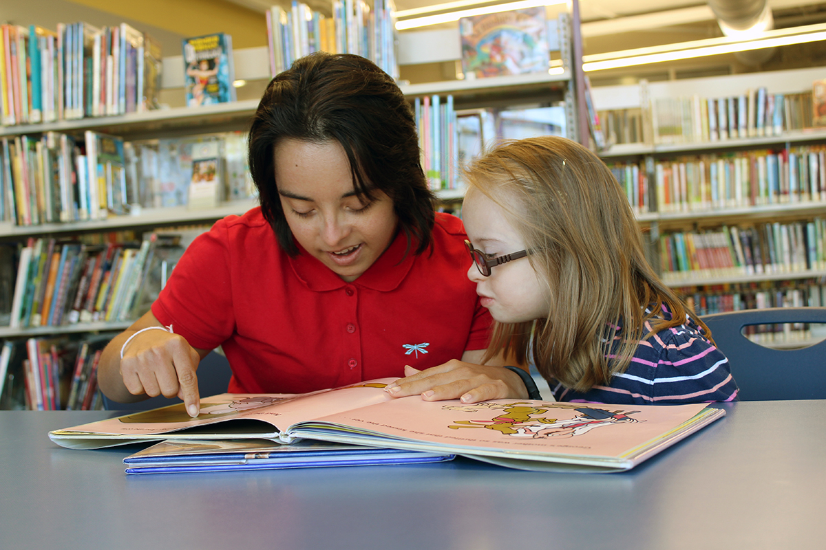 Photo of young woman with Down syndrome teaching reading to a child with Down syndrome.