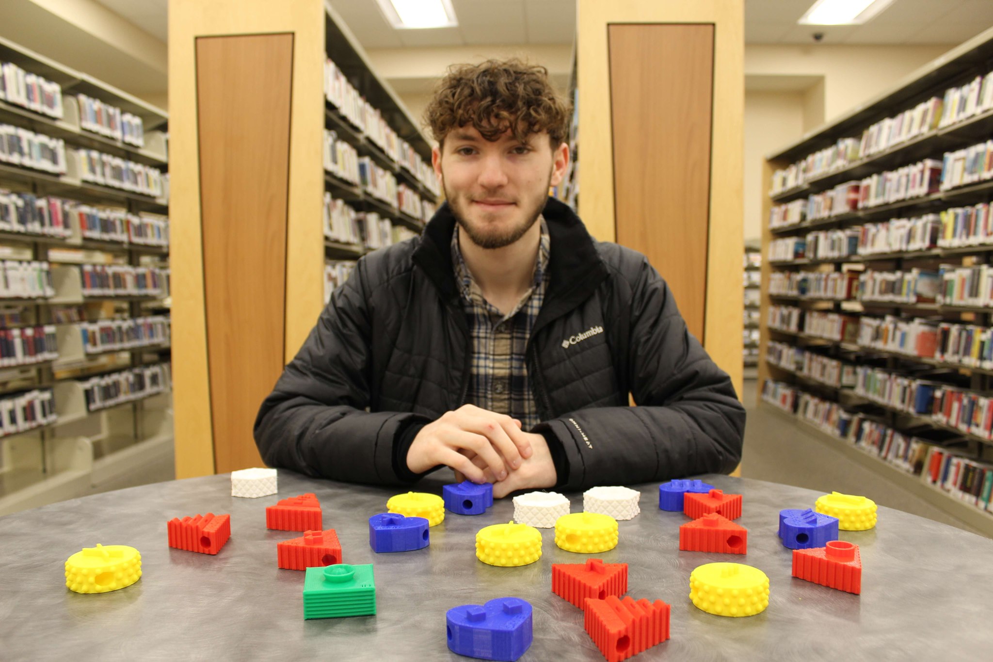 Young man with brown hair and beard wearing a black coat and brown flannel shirt seated at a library table in front of books. Multi-colored 3D printed objects are on the table.
