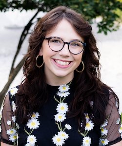 Mikaela Roark staff photo. She has long, curly, dark hair and is wearing a black blouse with daisies, black, rounded glasses, and gold hoop earrings.
