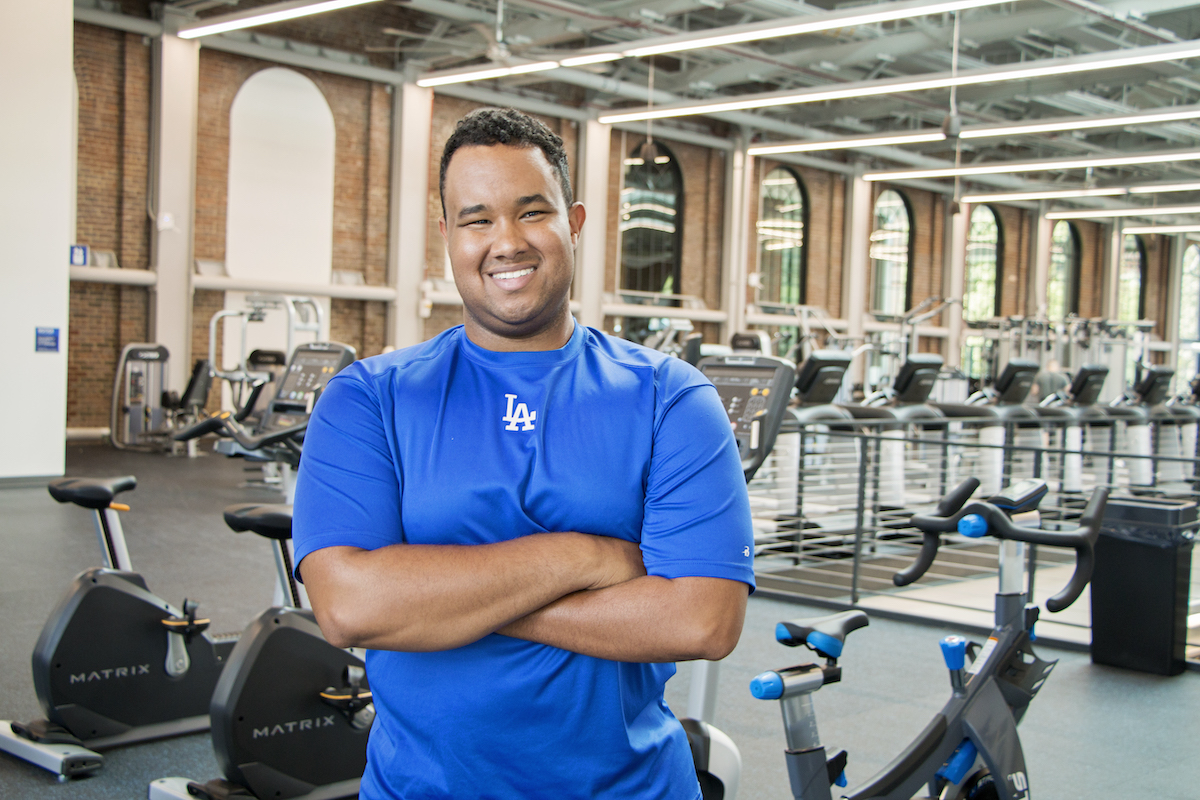 Morgan Turner wearing a UK blue t-shirt in a gym. He is smiling at the camera and has short, black hair.