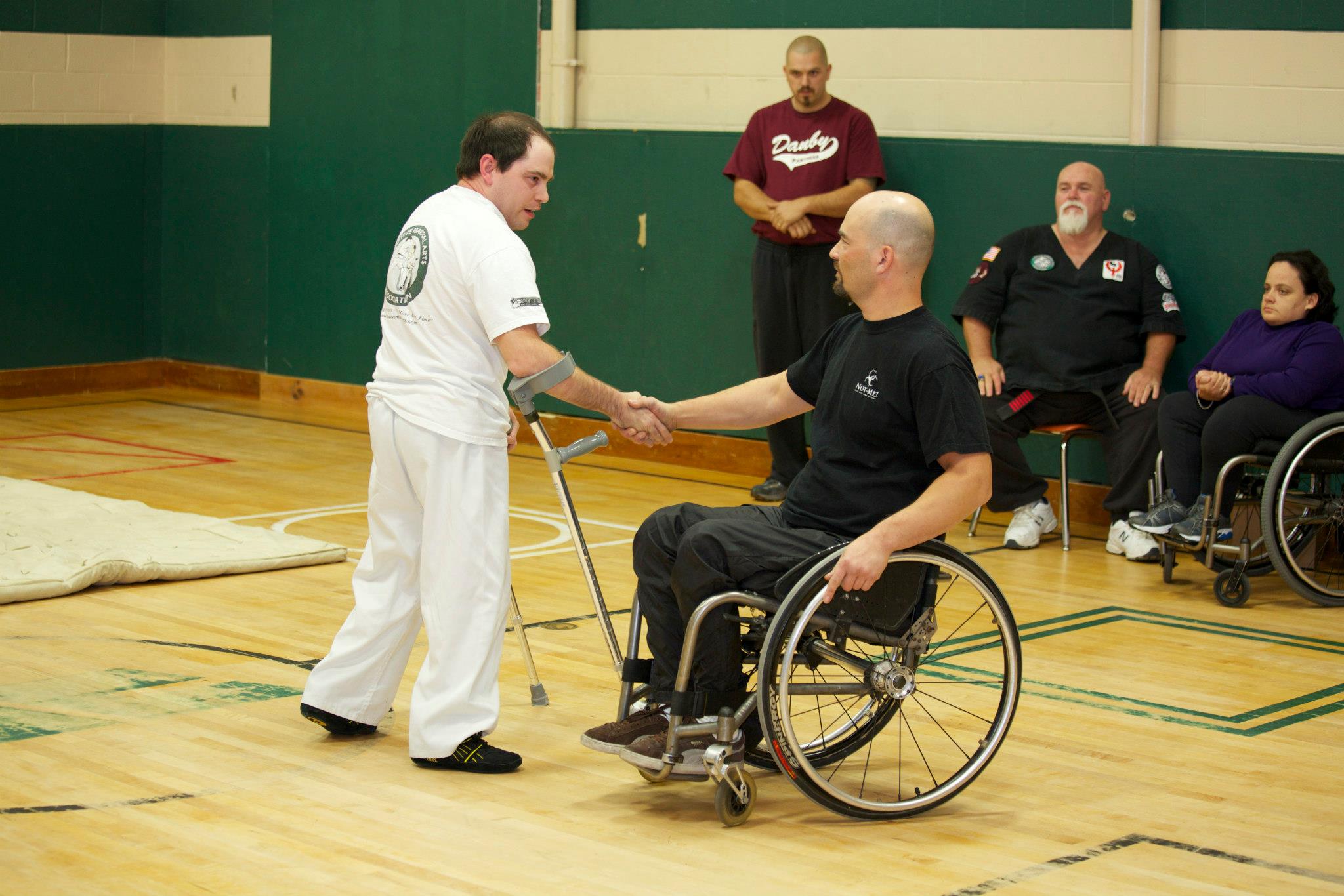 two male martial artists shaking hands; one man is wearing white and is standing and the other man is wearing black and uses a wheelchair for mobility. Three martial artists are shown in the background.