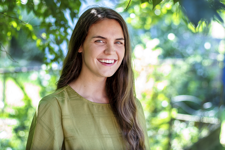 Austin wearing a green, linen shirt smiling in front of trees. She has long, brown wavy hair