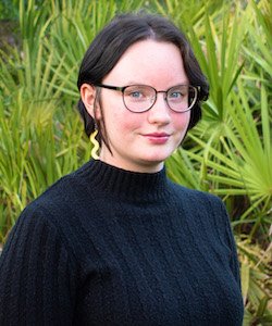 young woman wearing a black sweater, black glasses, and gold earrings standing in front of palm trees