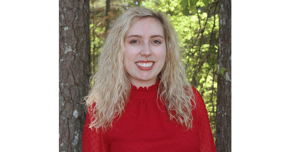 woman standing in front of trees is smiling has blonde hair and is wearing a red sweater