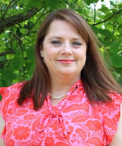 Paige Clements staff photo. She is wearing a red and pink paisley blouse and has shoulder-length, straight brown hair and is smiling at the camera