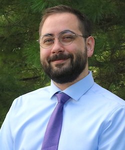 Benjamin Coffey staff photo. He has short, brown hair and a beard and is wearing glasses, a light blue button down and purple tie