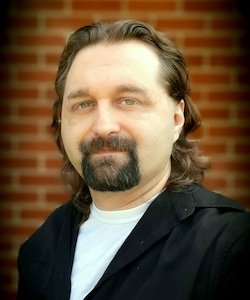 Derek Vincent staff photo. He is wearing a black suit and a white shirt on. He has long, dark brown hair and a beard and is looking at the camera. He is standing in front of a brick wall.