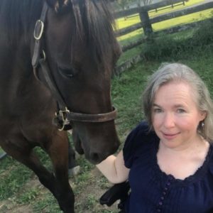 Woman with blond hair and fair skin is petting a dark brown horse that is standing in front of a black fence and green grass.