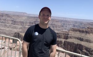 Stuart Rumrill standing in the Grand Canyon. He is wearing a black t-shirt and a baseball hat and smiling at the camera