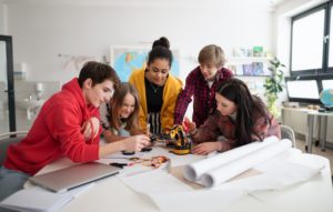 Five high school kids are gathered round a table collaborating on a robotics project. Wires and white rolled poster paper are visible on the table.