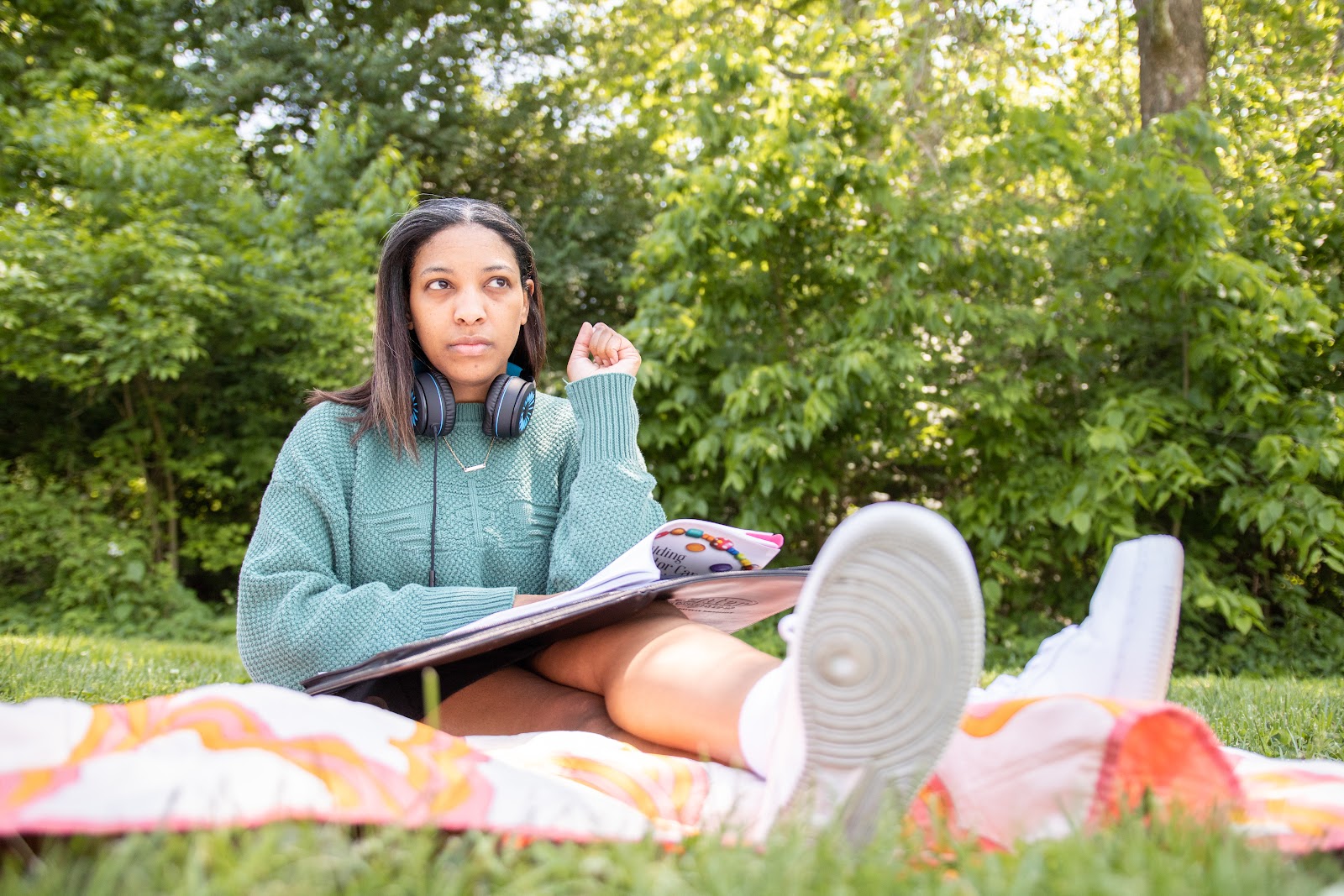 A young woman with headphones lowered to her neck sits on a blanket in a grassy outdoor space. She turns the page of a book and looks upward while contemplating. 